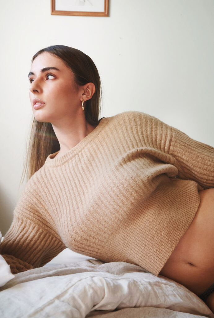 Marintia Bustos a model with long brown hair is seen in a relaxed pose on a bed wearing a beige sweater. She gazes upwards and a framed artwork hangs on the wall behind her.